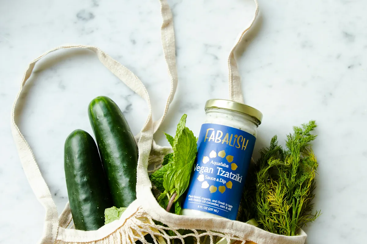 A mesh shopping bag with cucumbers, herbs, citrus and packaged groceries