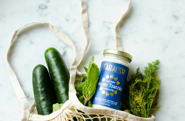 A mesh shopping bag with cucumbers, herbs, citrus and packaged groceries